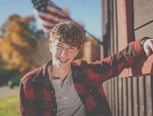 Teen boy with curly hair and braces smiling while leaning against a wooden wall, surrounded by autumn foliage and an American flag in the background, representing orthodontic treatment options for teens.